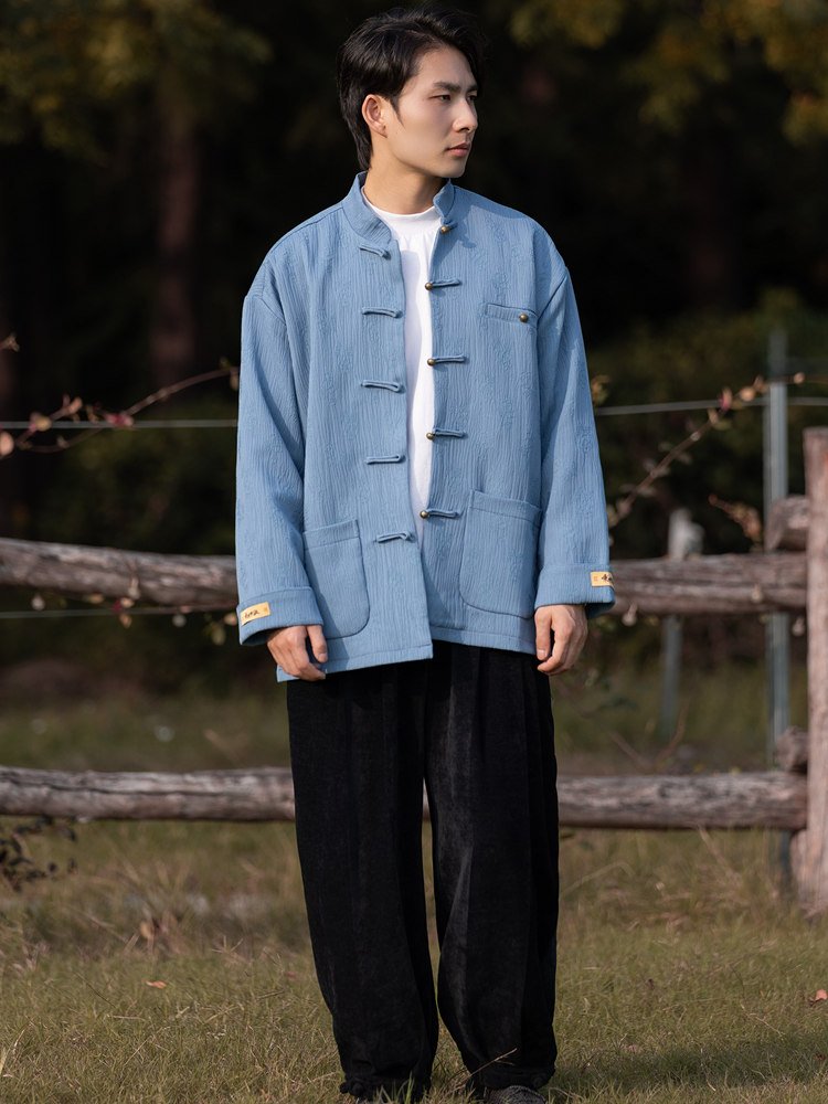 Full-body outdoor view of a man wearing a light blue Chinese Tang jacket with a mandarin collar and traditional frog buttons.